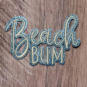 May include: A glittery blue embroidered patch featuring the text Beach Bum in a cursive and block font style. The lettering is outlined in a light beige thread, creating a layered effect against the sparkling blue background of the fabric patch.