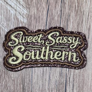 Peut inclure: Un écusson brodé avec un fond pailleté marron foncé et une bordure épaisse marron foncé. Le texte "Sweet, Sassy Southern" est cousu avec un fil de couleur crème clair dans une police cursive, centré sur l'écusson.