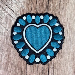 May include: A heart-shaped embroidered patch featuring a central teal heart outlined in white thread. The heart is surrounded by a border of alternating oval teal shapes and small white circular dots, all set against a black background.