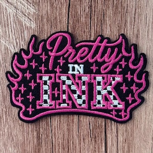 May include: A black embroidered patch featuring the pink text Pretty IN INK. The word INK is filled with a white and black checkered pattern. The design is surrounded by a pink flame border and small pink cross-shaped accents on a black background.