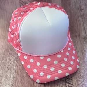 May include: A trucker hat featuring a solid white front panel and a pink mesh back with white polka dots. The curved brim is pink with white polka dots and a pink cord trim separates the brim from the front panel. A small pink button sits on top.