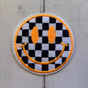 May include: A circular embroidered patch featuring a black and white checkered pattern. The design includes two oval-shaped orange eyes and a wide, curved orange smile. The entire circular edge is finished with a thick, bright orange border.
