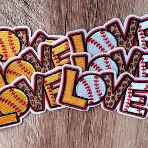 Puede incluir: Tres parches bordados con la palabra LOVE en letras de bloque. La L es amarilla, la O es una pelota de sóftbol amarilla o de béisbol blanca, la V tiene estampado de leopardo marrón y la E es blanca con costuras rojas. Borde blanco.