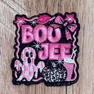 May include: A black embroidered patch featuring the pink text BOU JEE. The design includes a pink and white checkered ghost, a leopard print pumpkin, a pink bow, a pair of lips, and a pink drink cup, all surrounded by a scalloped black border.