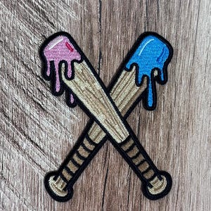Puede incluir: Un parche bordado que muestra dos bates de béisbol cruzados con un patrón de vetas de madera color canela. La parte superior de un bate está cubierta de glaseado rosa que gotea, mientras que el otro tiene glaseado azul, bordeado con hilo negro.