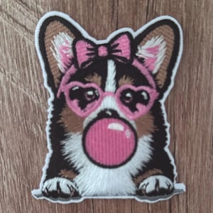 Może przedstawiać: Haftowana naszywka przedstawiająca trójkolorowego psa rasy Corgi z czarnym, brązowym i białym futrem. Pies ma na sobie różowe okulary w kształcie serc i pasującą różową kokardę na głowie, dmuchając przy tym dużą, okrągłą, jaskraworóżową bańkę z gumy.