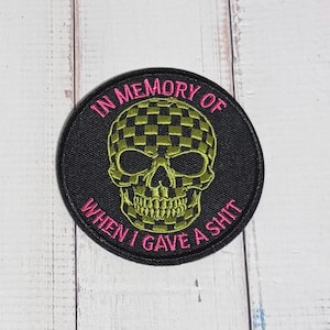 May include: A circular embroidered patch with a black background. It features a lime green checkered skull in the center. The text "IN MEMORY OF" is embroidered in pink at the top, and "WHEN I GAVE A SHIT" is embroidered in pink at the bottom.