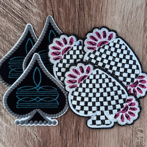 May include: A collection of embroidered spade-shaped patches. Some patches feature a black background with teal decorative line work, while others display a black and white checkered pattern accented with pink floral-like scalloped edges.