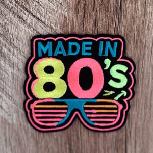 May include: A black embroidered patch featuring the text MADE IN in bright blue, the number 80's in lime green and hot pink, and a pair of shutter-style sunglasses in blue and pink with orange horizontal lines, all outlined in a thick black border.