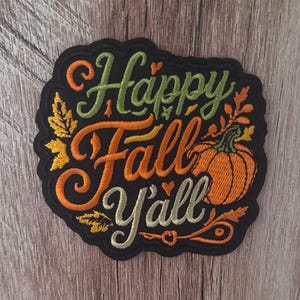 May include: A black embroidered patch featuring the text Happy Fall Y'all in green, orange, and cream thread. The design includes an orange pumpkin, small hearts, and autumn leaves in yellow and orange, all set against a black background.
