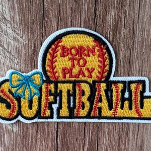 May include: An embroidered patch featuring a yellow softball with red stitching and the text BORN TO PLAY in red. Below, the word SOFTBALL is written in yellow with red stitching details, accented by a small blue bow on the left side.