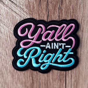May include: A black embroidered patch featuring the text Y'all Ain't Right. The word Y'all is stitched in pink, the word AIN'T is in white, and the word Right is in light blue. The patch has a black border and a textured, raised thread finish.