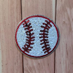 Puede incluir: Un parche circular de béisbol con lentejuelas blancas que cubren la superficie principal, acentuado por dos líneas curvas de lentejuelas rojas que representan las costuras. El parche tiene un borde rojo fino y un borde exterior de tela brillante plateada.