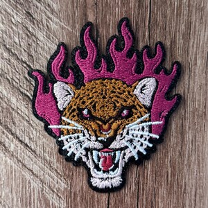 May include: An embroidered patch featuring a leopard head with a snarling expression, bared teeth, and a red tongue. The leopard is surrounded by stylised magenta flames with a black border. The design includes white whiskers and ears.