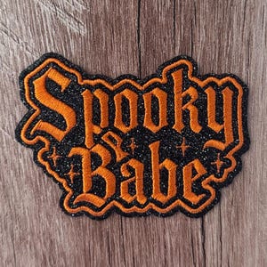 May include: An embroidered patch featuring the text Spooky Babe in orange gothic-style lettering. The background of the patch is black with a glittery texture, and the entire design is outlined in a matching orange border with small star accents.