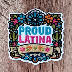 Könnte beinhalten: Ein bestickter Aufnäher mit dem Text PROUD LATINA in blauen und rosa Buchstaben. Das Design ist von einem bunten Blumenrand mit grünen Kakteen, orangefarbenen, blauen und rosa Blüten sowie einem beigen Banner unten umgeben.