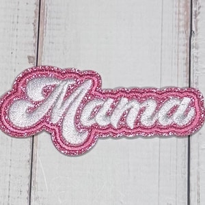 May include: An embroidered patch featuring the word Mama in a cursive script font. The lettering is rendered in a shimmering silver thread, while the surrounding border and background layer consist of a vibrant, glittery pink fabric texture.