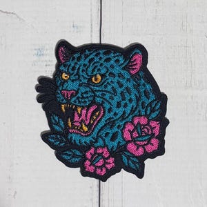 May include: An embroidered patch featuring a teal leopard head with black spots, yellow eyes, and an open mouth showing sharp teeth. The design includes bright pink accents on the ears, nose, and mouth, surrounded by pink flowers and green leaves.