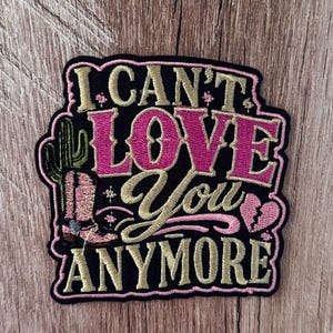 Könnte beinhalten: Ein schwarzer bestickter Aufnäher mit dem Text I CAN'T LOVE YOU ANYMORE in goldenem und rosa Garn. Das Design enthält einen grünen Kaktus, einen rosa Cowboystiefel und ein kleines gebrochenes Herz, alles mit einem rosa Rand auf schwarzem Grund.