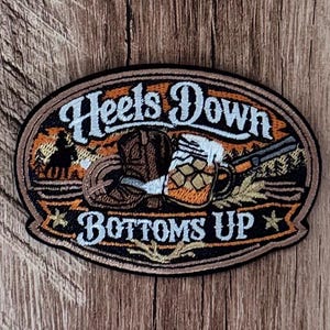 May include: An oval embroidered patch featuring the text "Heels Down" and "Bottoms Up" in white lettering. The design includes a brown cowboy boot, a glass mug filled with a golden beverage, and a rifle, set against an orange and black background.