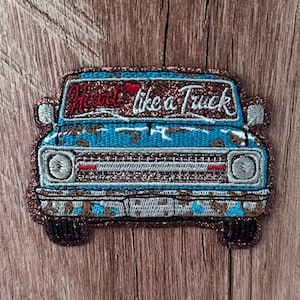 May include: An embroidered patch shaped like the front of a vintage truck. The truck is blue with brown distressed details and a glittery copper border. The windshield features the red and white embroidered text Heart like a Truck.