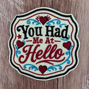 May include: A shield-shaped embroidered patch with a cream background and a black border. It features the text "You Had Me At Hello" in black and red script. The design is decorated with four red hearts and light blue swirling flourishes.