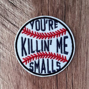 Könnte beinhalten: Ein runder, bestickter Aufnäher in Form eines Baseballs mit weißem Hintergrund, schwarzem Rand und roten Nahtdetails. Der Text "YOU'RE KILLIN' ME SMALLS" ist in schwarzer Schrift in der Mitte des Baseball-Motiv-Aufnähers eingestickt.