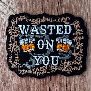 May include: A rectangular black embroidered patch featuring the white text "WASTED ON YOU" with two illustrations of amber-colored whiskey glasses. The patch is bordered by a decorative, intricate tan-colored vine-like pattern on a black background.