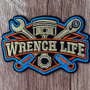 May include: An embroidered patch featuring a central engine piston flanked by a wrench and a ratchet. The design includes orange and light blue borders with the text WRENCH LIFE in beige lettering against a black background.