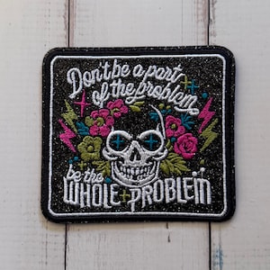 Könnte beinhalten: Ein quadratischer Stickaufnäher mit schwarzem Glitzerhintergrund, der einen weißen Totenkopf mit blauen Kreuzaugen zeigt, umgeben von rosa und grünen Blumen und Blitzen, mit dem Text Don't be a part of the problem be the WHOLE PROBLEM.