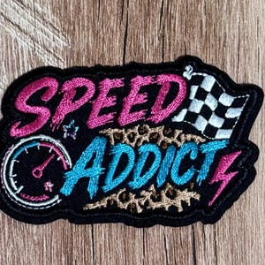 May include: An embroidered patch with a black border featuring the pink text SPEED and blue text ADDICT. The design includes a black and white checkered flag, a speedometer, a small star, a pink lightning bolt, and a brown leopard print pattern.