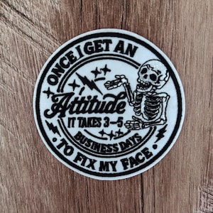 May include: A circular embroidered patch featuring a black and white design of a skeleton. The text reads: ONCE I GET AN Attitude IT TAKES 3-5 BUSINESS DAYS TO FIX MY FACE. The patch includes graphic elements of lightning bolts and stars.