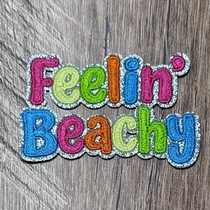 May include: An embroidered patch featuring the text Feelin Beachy in two rows. The letters are stitched in vibrant shades of pink, lime green, orange, and blue, all outlined with a shimmering silver border that follows the shape of the words.
