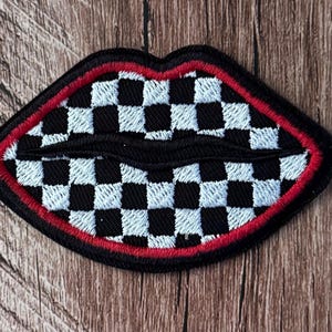 Könnte beinhalten: Ein gestickter Aufnäher in Lippenform mit einem schwarz-weißen Schachbrettmuster. Die Lippen sind mit einem dünnen roten Rand und einer dicken schwarzen Außenkante versehen, was eine markante Form erzeugt, die die detaillierte Stickerei betont.