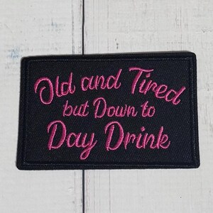 May include: A rectangular black embroidered patch featuring the pink stitched text Old and Tired but Down to Day Drink. The patch has a thick black border and a textured fabric surface, with the lettering displayed in a cursive script font.