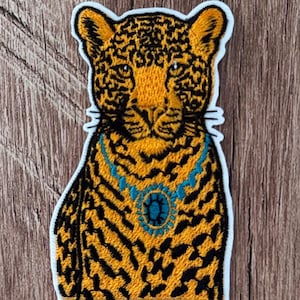 May include: An embroidered iron-on patch featuring a leopard with yellow fur and black spots. The leopard is wearing a bright turquoise necklace with a central oval-shaped blue gemstone. The patch has a thick white border and a flat design.