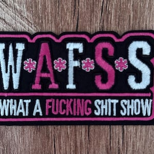 Puede incluir: Un parche bordado rectangular con fondo negro y borde rosa. El texto W*A*F*S se muestra en blanco y rosa con iconos de flores rosas. Debajo, el texto WHAT A FUCKING SHIT SHOW está bordado en blanco y rosa.