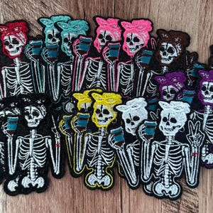 May include: A collection of embroidered patches featuring skeletons wearing headscarves in various colors, including red, pink, yellow, white, and purple. Each skeleton holds a blue cup and displays a peace sign with one hand.
