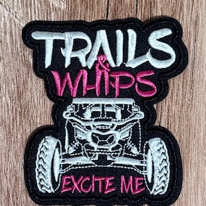 Puede incluir: Un parche bordado negro con el texto blanco TRAILS, el texto rosa & WHIPS y el texto rosa EXCITE ME. El diseño incluye un gráfico bordado en blanco de la vista frontal de un vehículo todoterreno con neumáticos grandes.