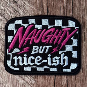 May include: A rectangular embroidered patch featuring a black and white checkered border. The text "NAUGHTY" is stitched in bright pink, followed by "BUT" and "nice-ish" in white, with two small pink lightning bolt icons between the words.