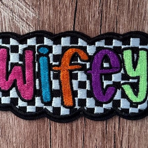 May include: An embroidered patch featuring the word wifey in multi-colored letters including pink, blue, orange, purple, and lime green. The text is set against a black and white checkered background with a thick black stitched border outline.
