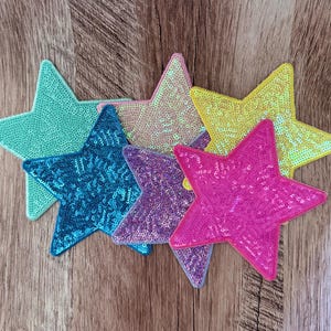 May include: A collection of five star-shaped patches covered in shimmering sequins. The set includes individual stars in bright shades of mint green, light pink, deep blue, purple, and yellow, each featuring a dense, reflective sequin texture.