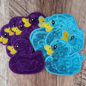 Op de afbeelding: Een verzameling eendvormige patches bedekt met glinsterende lovertjes. De patches zijn gerangschikt in twee afzonderlijke groepen, waarbij de ene set is voorzien van dieppaarse lovertjes en de andere set van helder turquoise lovertjes, beide met gele snavels.