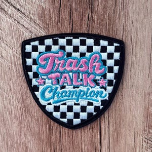 May include: A shield-shaped embroidered patch featuring a black and white checkered pattern. The text "Trash TALK Champion" is stitched in pink and blue cursive and block lettering, accented by two small pink stars on the sides of the word TALK.