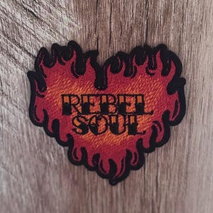 Puede incluir: Un parche bordado con forma de corazón rodeado de llamas. El corazón presenta un degradado de hilos rojos y naranjas con un borde negro grueso. Las palabras REBEL SOUL están cosidas en letras negras en negrita en el centro del corazón.