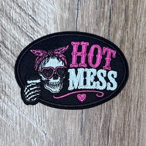 May include: An oval-shaped embroidered patch with a black background. It features a skull wearing a pink bandana and heart-shaped sunglasses, holding a cup. The text reads HOT in pink and MESS in light blue, with a small pink heart below the text.