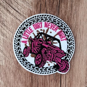 May include: A circular embroidered patch featuring a pink off-road vehicle on a white background with brown mud splatters. The text A LITTLE DIRT NEVER HURT is stitched in pink. The border is a black and white tire tread pattern design.