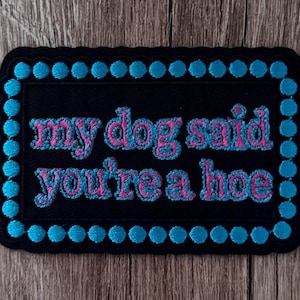 May include: A rectangular black embroidered patch with a scalloped edge featuring a border of bright blue dots. The centre displays the text "my dog said you're a hoe" stitched in pink letters with a bright blue outline on a black background.