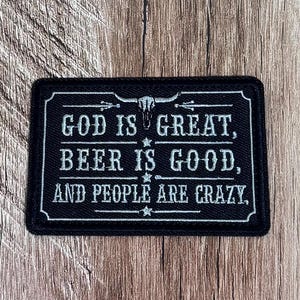 May include: A rectangular black embroidered patch featuring a white steer skull icon at the top and the text "GOD IS GREAT, BEER IS GOOD, AND PEOPLE ARE CRAZY." The design includes thin horizontal lines and small star accents within a decorative border.