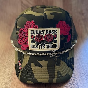 May include: A camouflage trucker hat featuring a rectangular patch with the text "EVERY ROSE HAS ITS THORN" and red rose illustrations. The hat is accented with a silver-toned barbed wire chain across the brim and three small red circular studs.